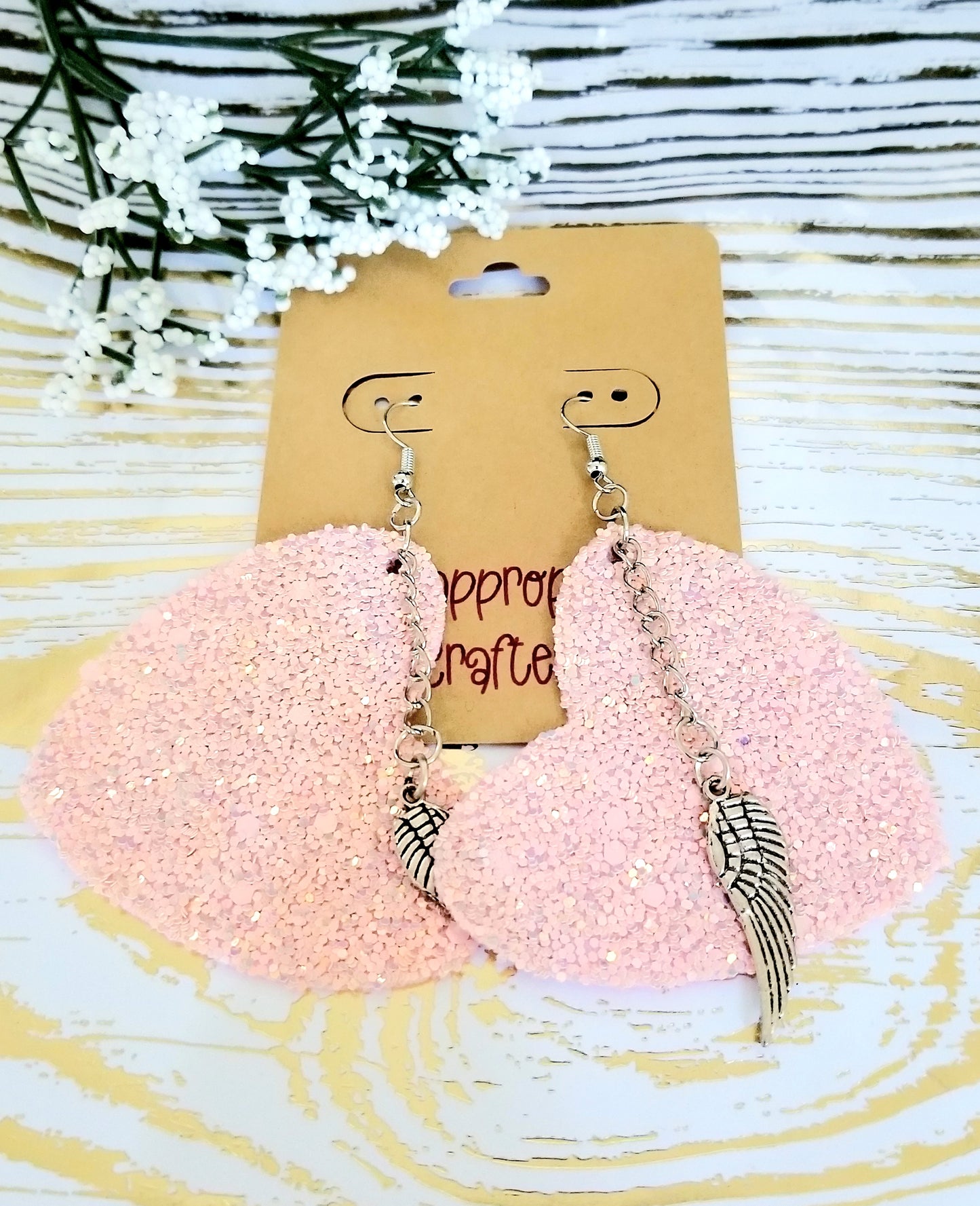 A pair of pink glittery heart shaped earrings with silver chains and wing charms, displayed on a card.