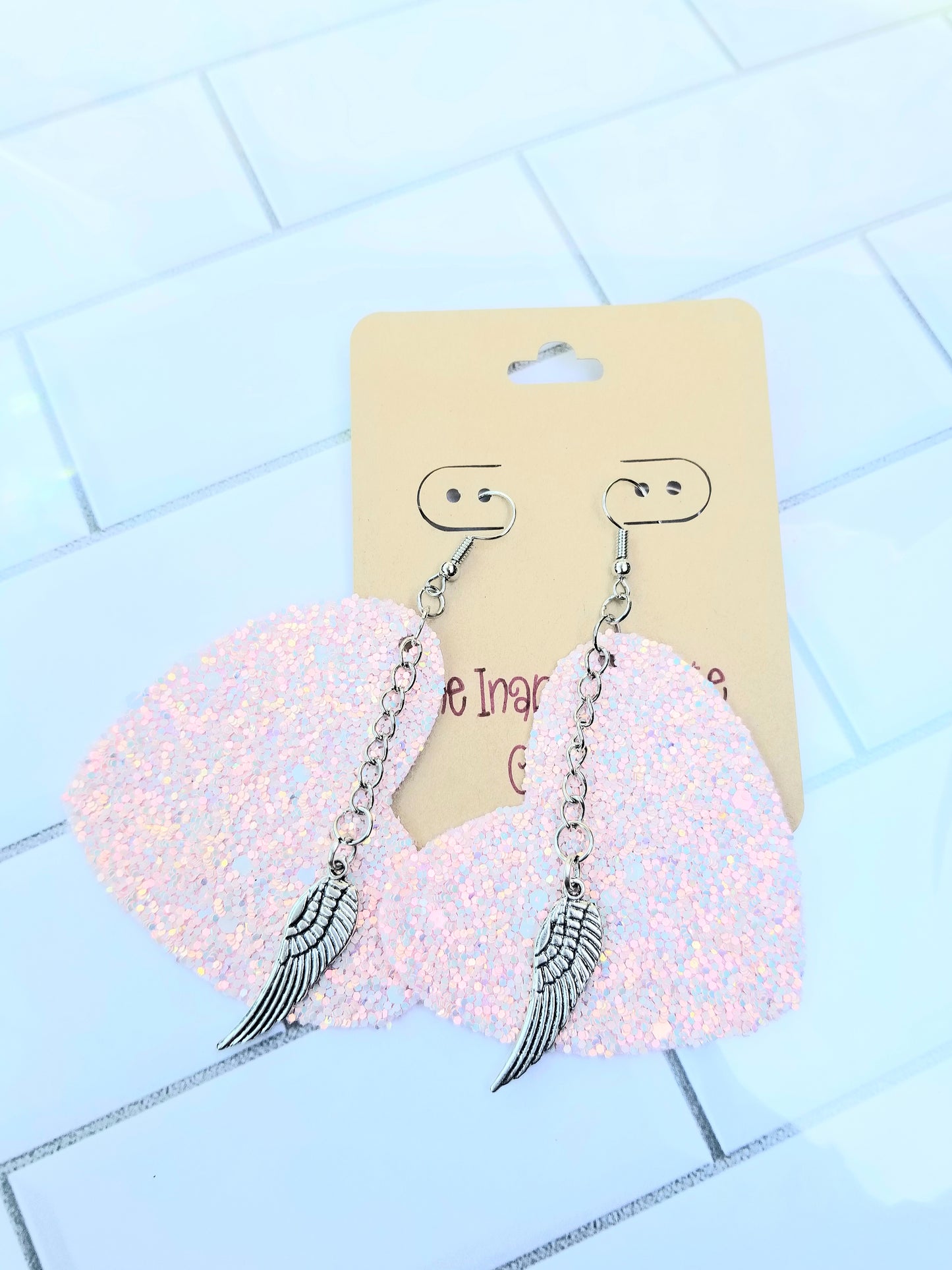 A pair of pink glittery heart shaped earrings with silver chains and wing charms, displayed on a card.
