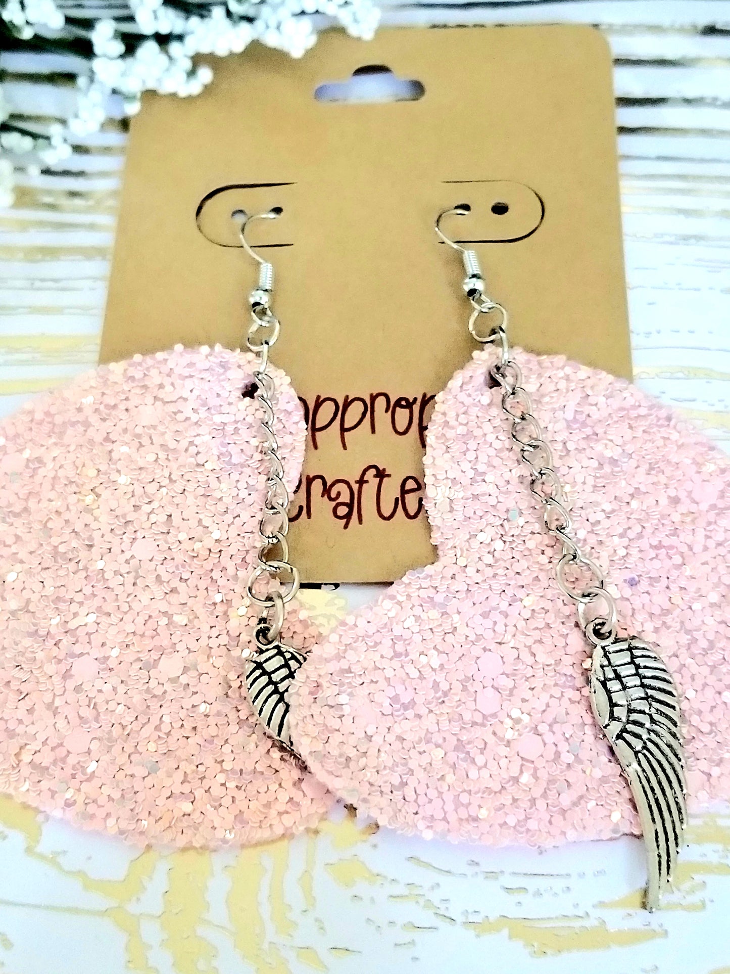 A pair of pink glittery heart shaped earrings with silver chains and wing charms, displayed on a card.
