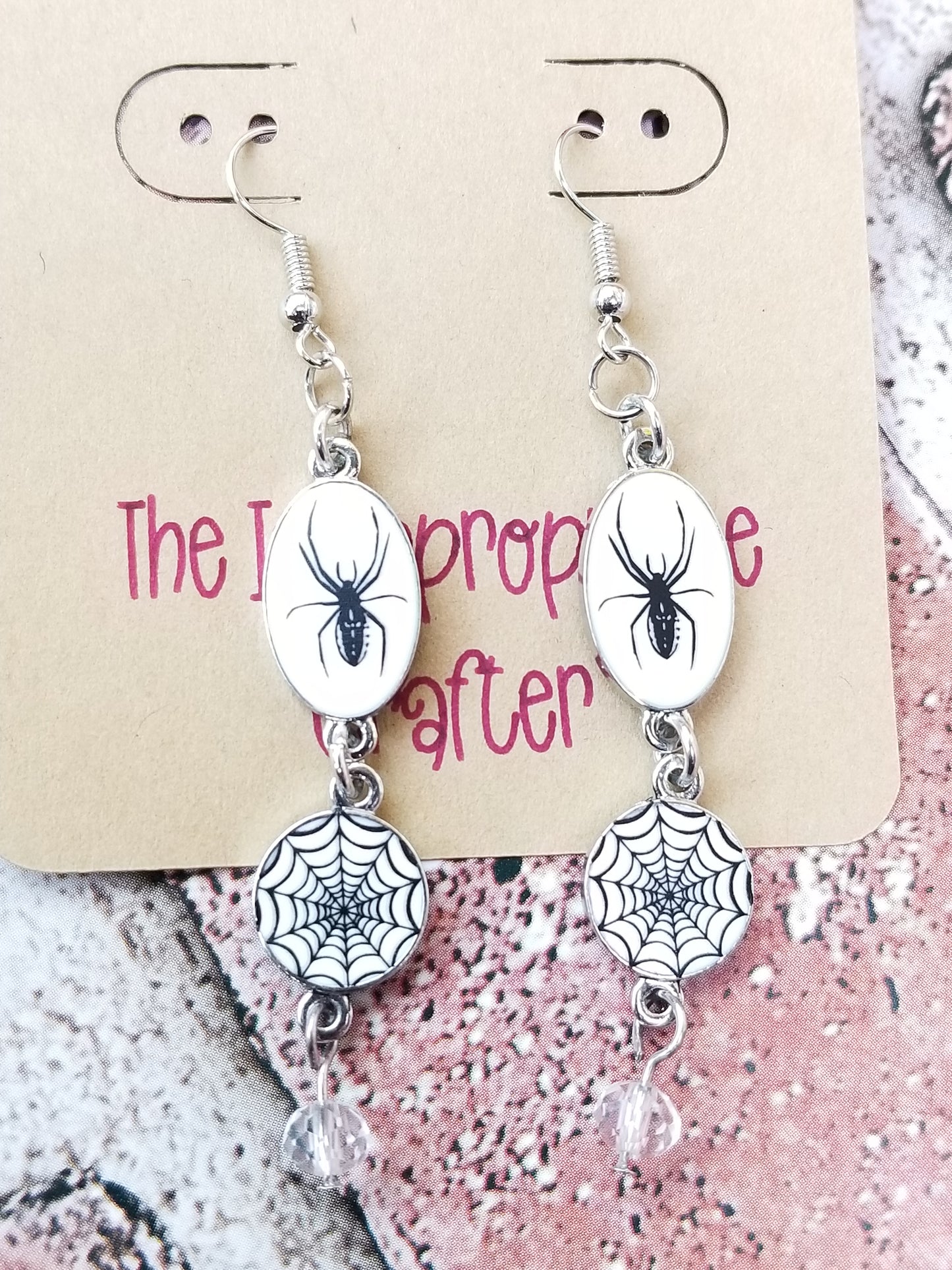 Silver colored dangle earrings featuring a spider and web design hanging on a card.