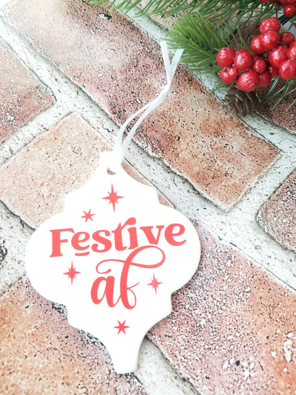A ceramic ornament with a red 'Festive AF' design, hanging against a brick wall background with holly and berries.