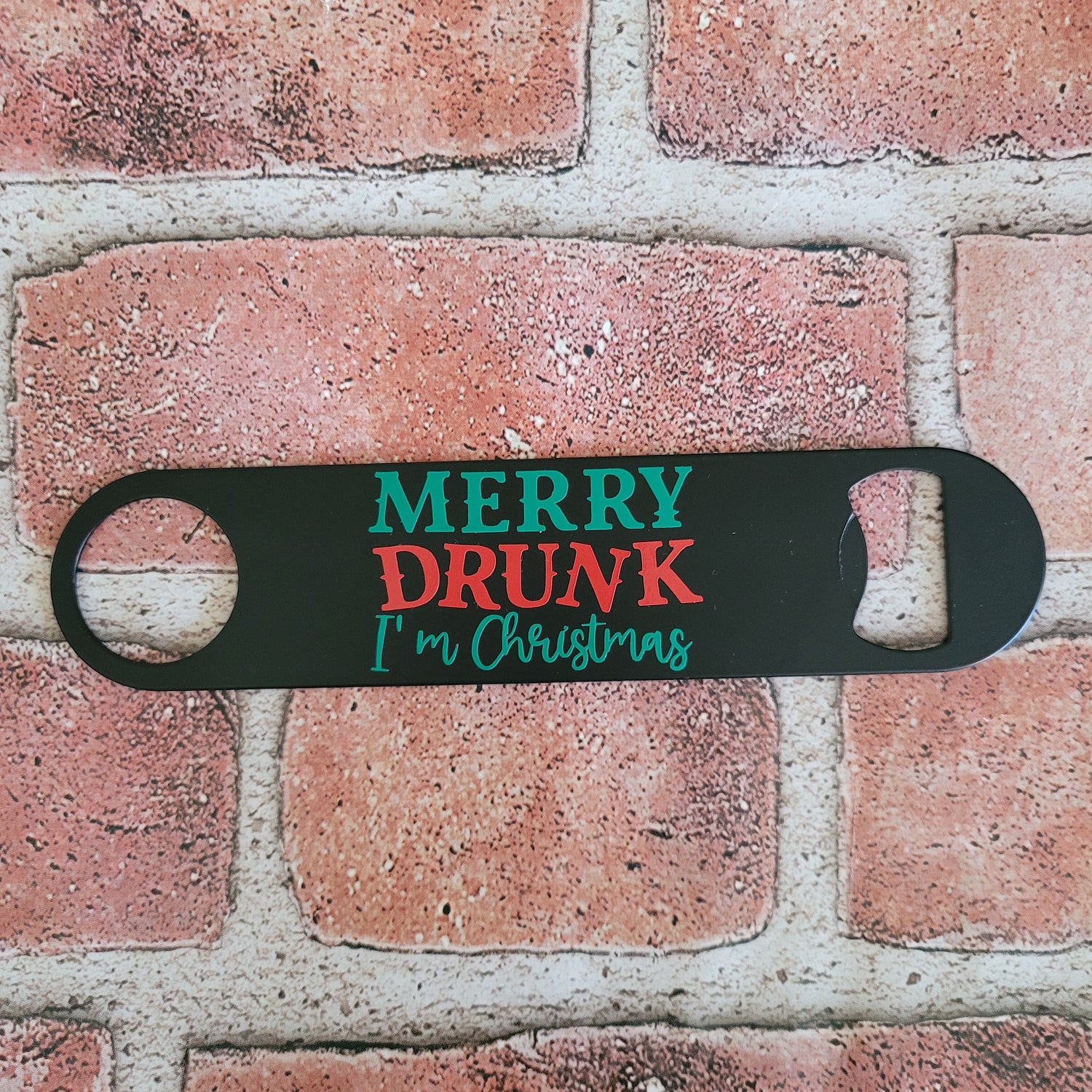 Black bottle opener with 'Merry Drunk I'm Christmas' text on a brick wall background
