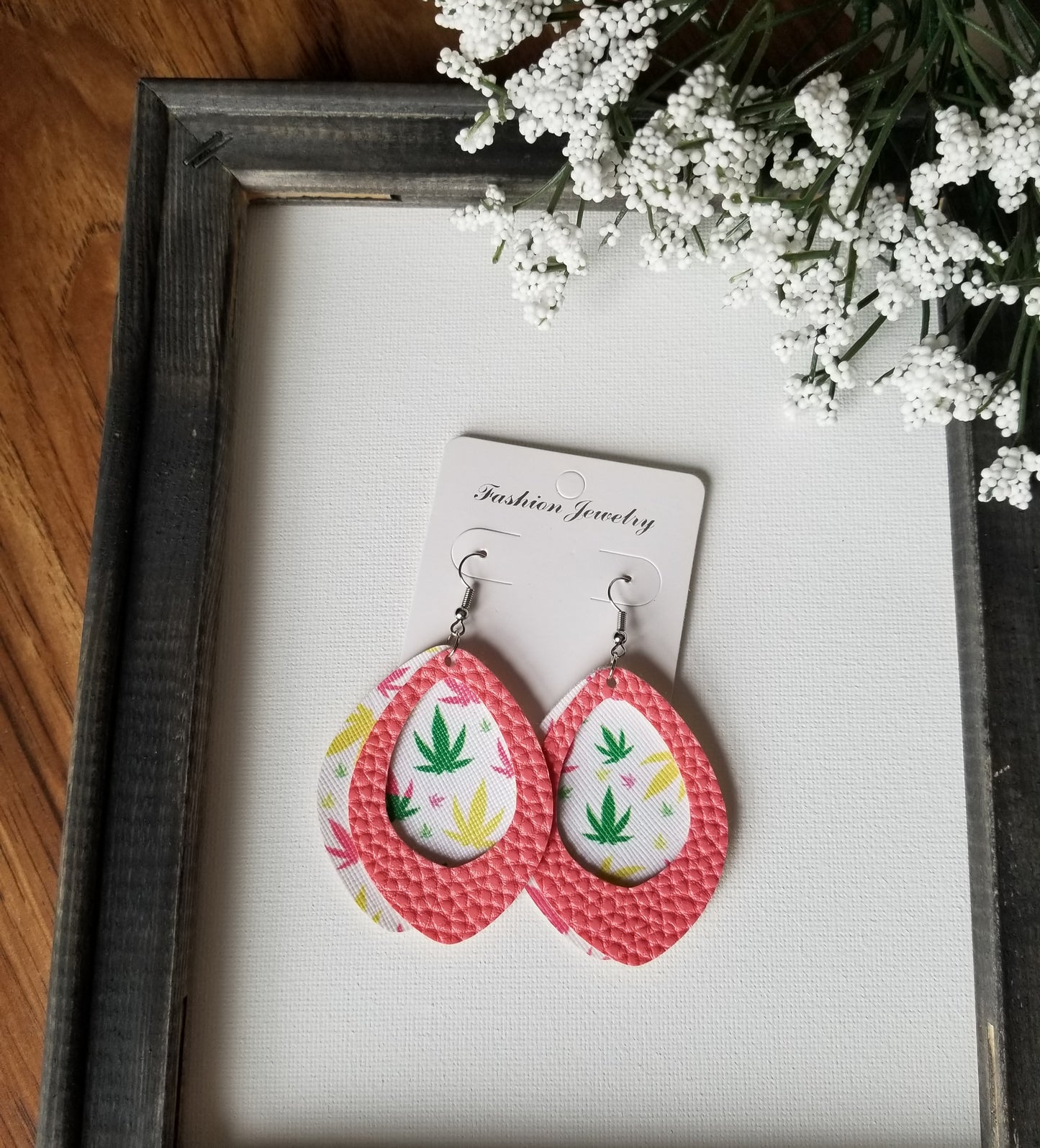 A pair of faux leather earrings. The top layer is coral pink and the bottom layer has a multicolored marijuana design.  The earrings are on a product card displayed on a frame.