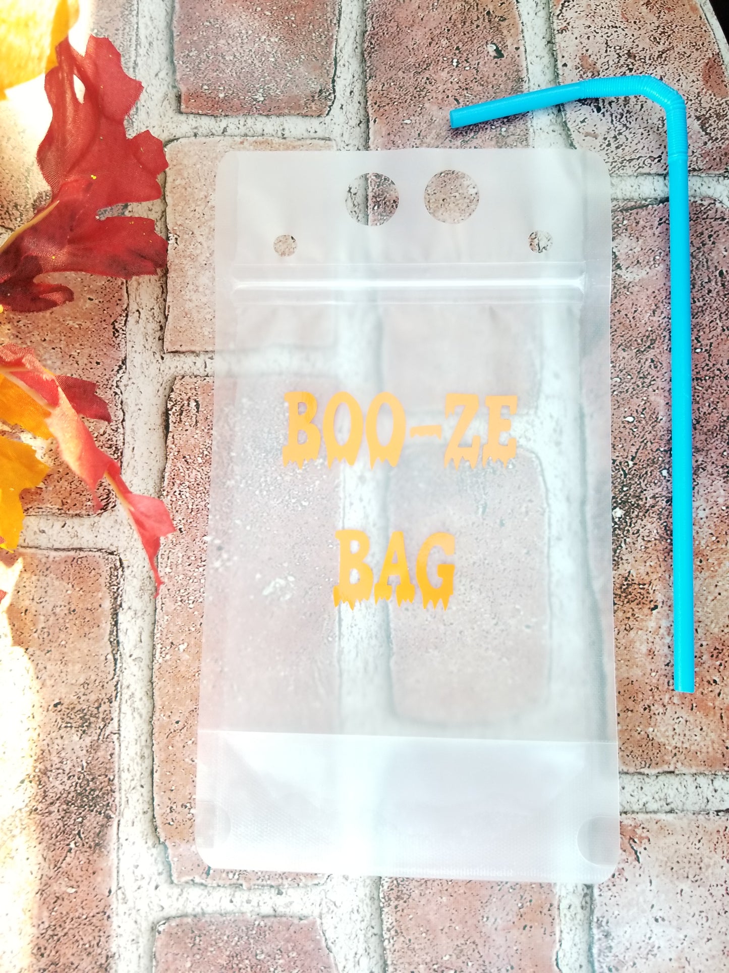 A clear plastic drink pouch with orange vinyl design, labeled 'BOO-ZE BAG', displayed on a brick surface with autumn leaves around.