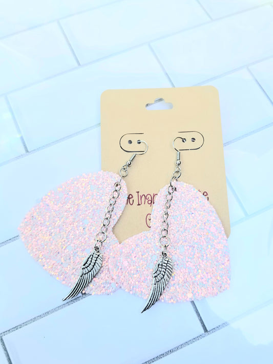 A pair of pink glittery heart shaped earrings with silver chains and wing charms, displayed on a card.