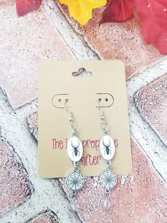 A pair of silver-colored earrings featuring spider and web designs, with a clear faceted bead, displayed on a card.
