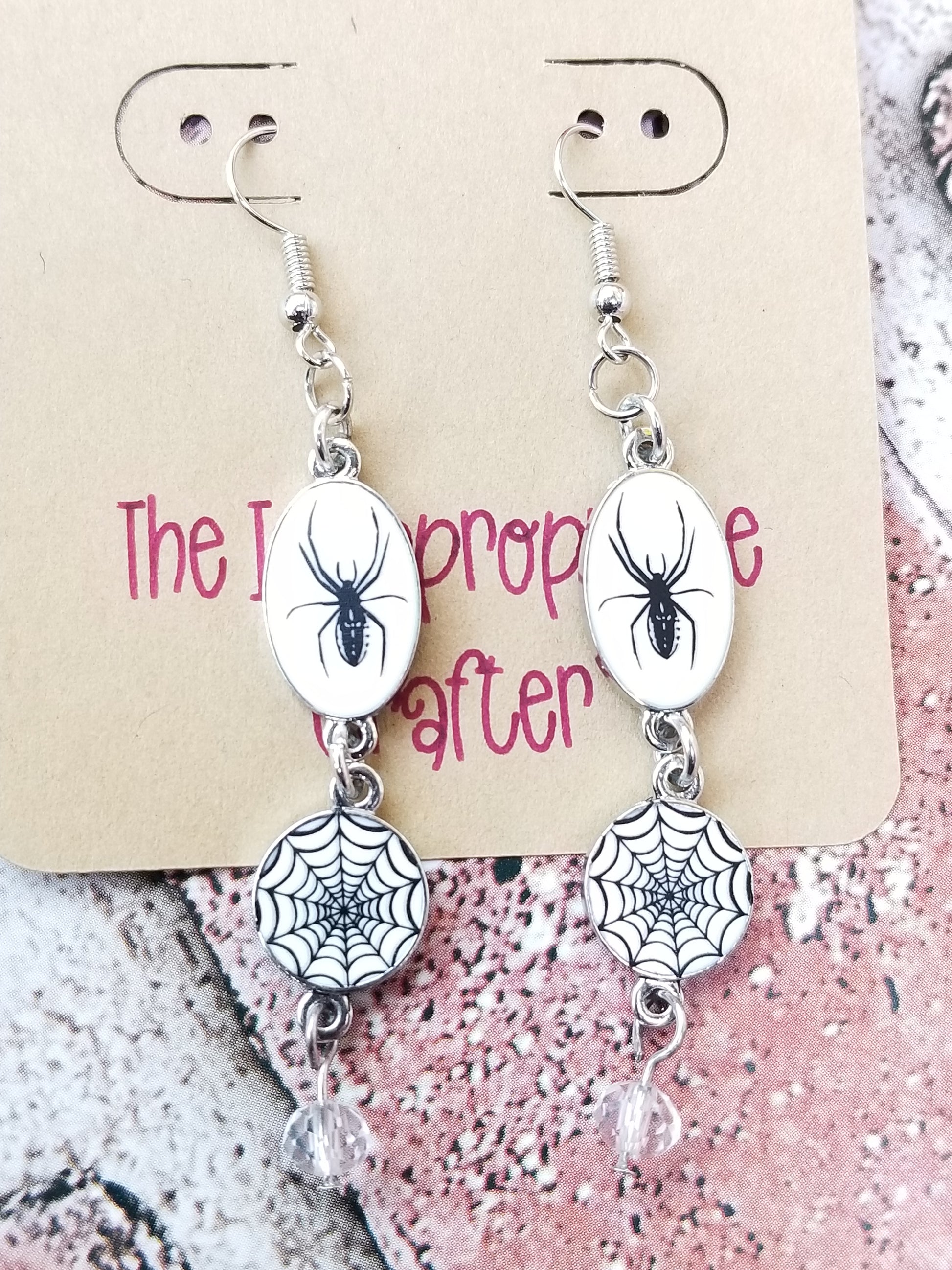 Silver colored dangle earrings featuring a spider and web design hanging on a card. 